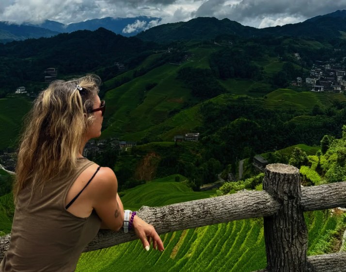 A visitor leaning on a timber rail at a Longji Rice Terraces viewpoint looking across the valley to terraced slopes